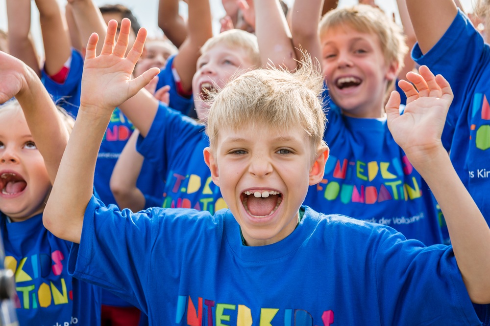 Kinder in blauen T-Shirts lachen und jubeln mit erhobenen Händen. Fröhliche Stimmung bei einem Outdoor-Event.